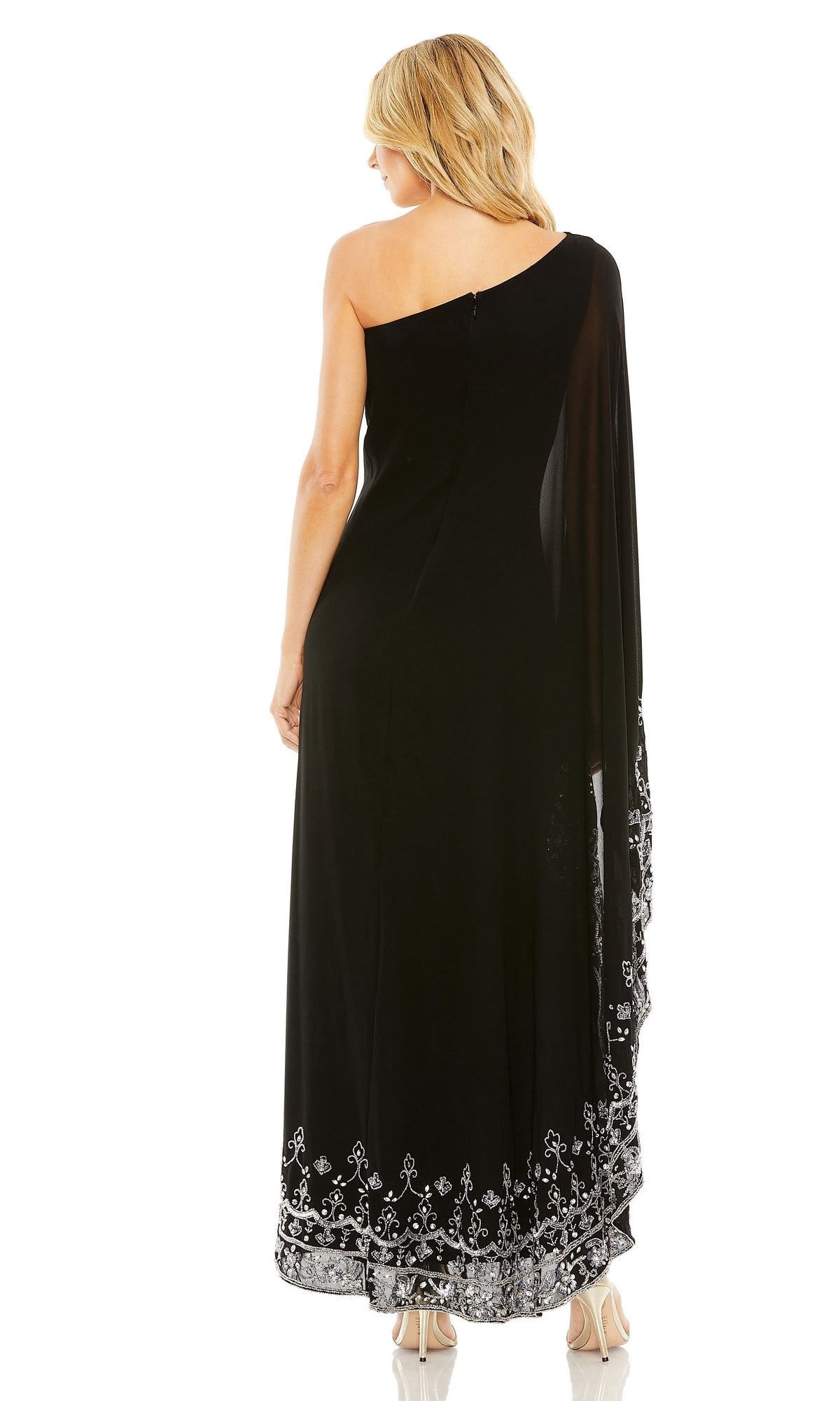 A woman faces away, wearing the Long Formal Dress 9251 by Mac Duggal—a one-shoulder black gown with a long sleeve cape and silver beaded trim—paired with light-colored high heels.